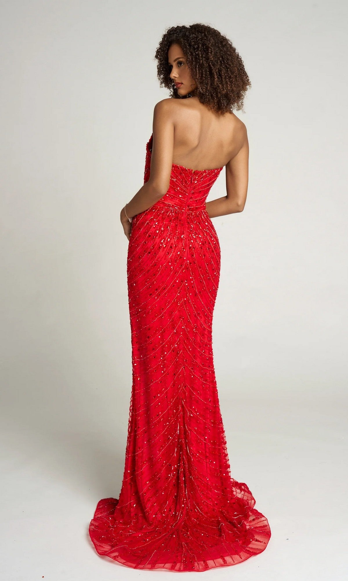 A woman stands facing away, wearing the Portia and Scarlett PS26421C long formal dress in red—a strapless, beaded prom gown with floor-length elegance—set against a plain light background.